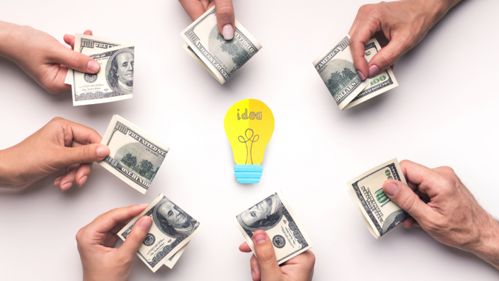 Siete manos sostienen fajos de billetes de 100 dólares estadounidenses, acercándolos hacia el centro, donde hay una figura de un foco amarillo con la palabra "idea" escrita en él. La imagen representa la inversión en una idea o negocio mediante un sistema colaborativo, similar a las tandas, donde varias personas aportan dinero con el fin de financiar un proyecto o recibir un fondo en determinado turno.