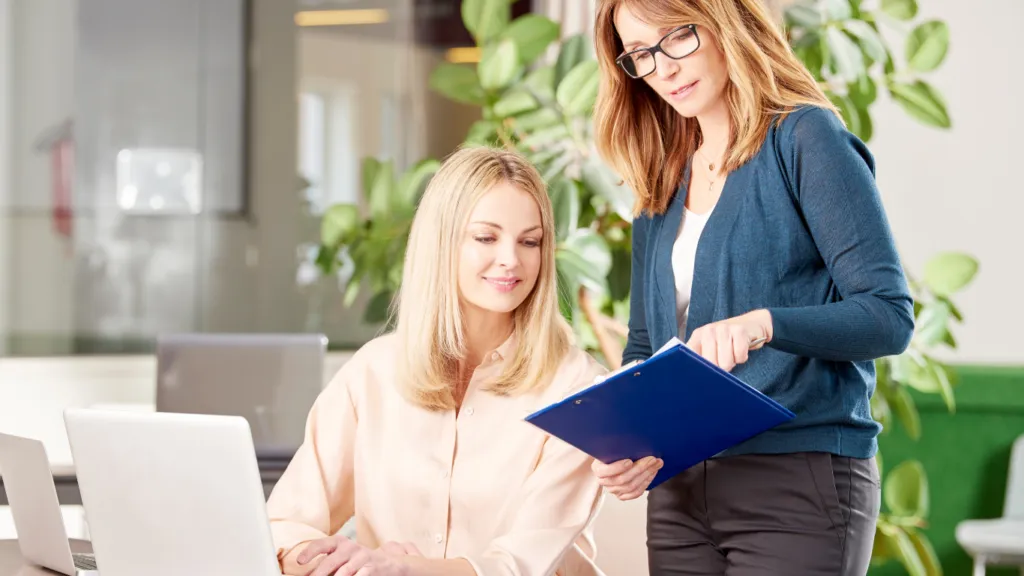 Dos mujeres en un entorno de oficina colaborativo. Una mujer de cabello rubio está sentada frente a una laptop, sonriendo, mientras la otra, con gafas y carpeta azul en mano, le explica algo