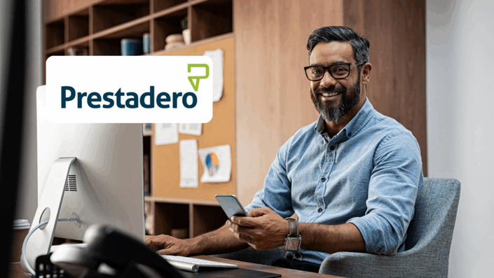 Hombre de barba y gafas sonriente trabajando en su escritorio, sosteniendo un celular en una mano y mirando hacia la cámara. A la izquierda, se muestra el logo de Prestadero.