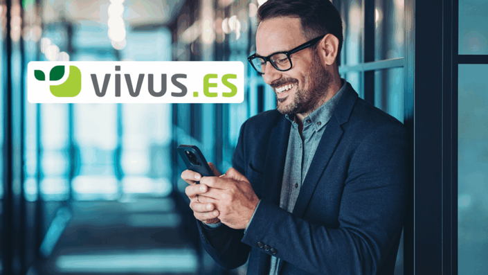 Hombre elegante sonriendo mientras usa su celular en un pasillo de oficinas, con el logotipo de Vivus.es representando préstamos personales fáciles desde el móvil.