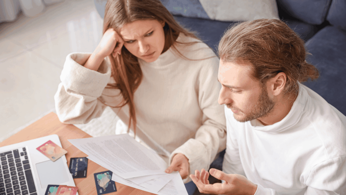 Una pareja sentada en una mesa, el hombre usando una calculadora y la mujer escribiendo en un documento, con un teléfono celular y una laptop cerca, representando la revisión de estados de cuenta para identificar cargos no reconocidos en tarjeta.
