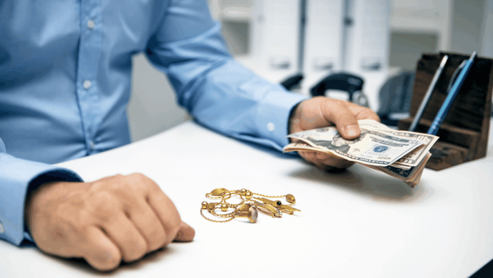 Hombre con camisa azul sosteniendo dinero en efectivo sobre una mesa blanca con joyas de oro, ilustrando el proceso de empeñar oro.