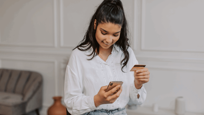 Joven sonriente usando su smartphone y sosteniendo una de sus primeras Tarjetas para principiantes, realizando una compra online.