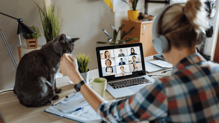 Persona trabajando en casa con auriculares, participando en una videollamada en su laptop, con un gato sentado en la mesa.