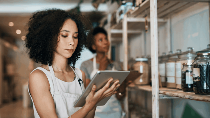 Mujer joven con delantal usando una tablet en una tienda moderna, gestionando inventario digital junto a estantes con frascos.