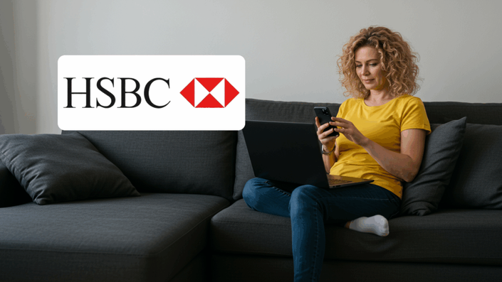 Mujer rubia de cabello rizado, sentada en un sofá gris, usando su celular mientras tiene una computadora portátil en las piernas. A la izquierda aparece el logotipo del banco HSBC.